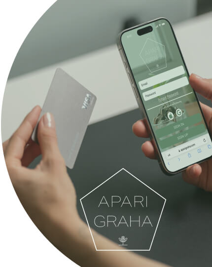 A person holds a mobile phone with the Aparigraha app home screen in one hand and a credit card in the other.